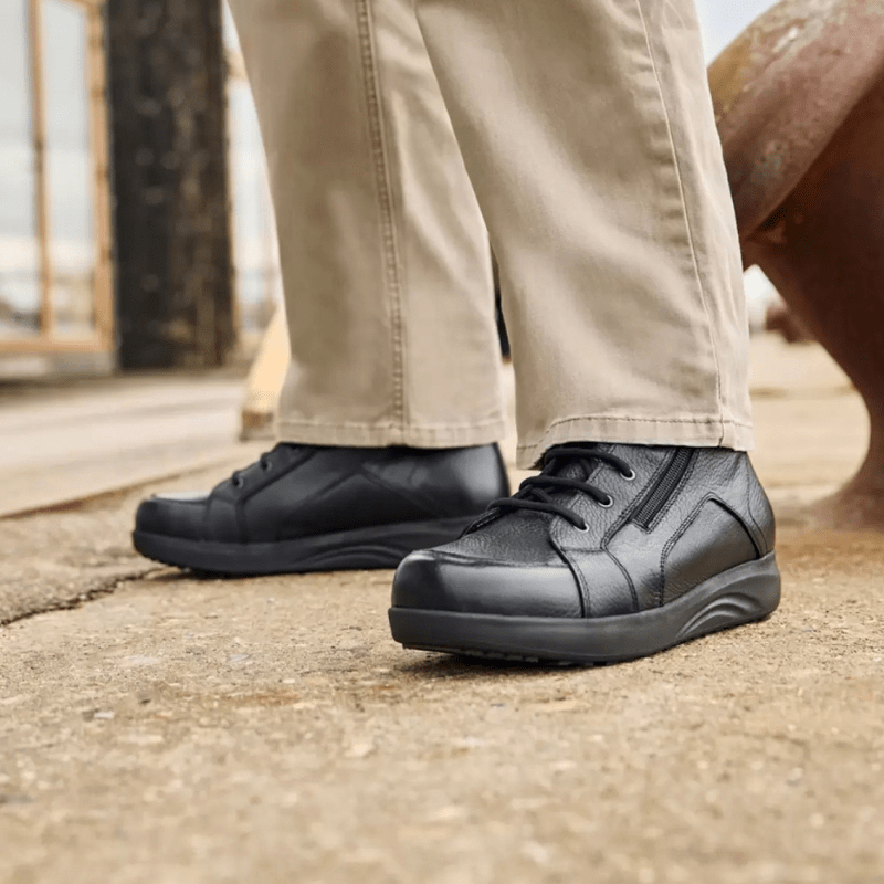 Un hombre con pantalones beige y botas New Feet negras de hombre de pie sobre una superficie gris