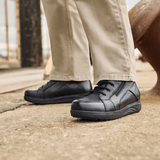 Un hombre con pantalones beige y botas New Feet negras de hombre de pie sobre una superficie gris