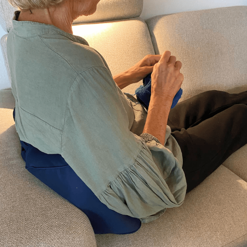Persona mayor sentada en un sofá con un cojín lumbar azul para apoyo de espalda, tejiendo y relajándose